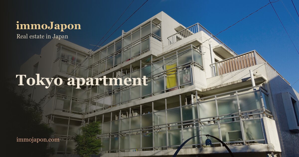 A four-storey apartment block in Shinjuku ward, Tokyo: white facade, open walkways and balconies behind glass railings, laundry and bicycles visible, trees at the foot of the building under a blue sky, the kind of condominium bought in the city