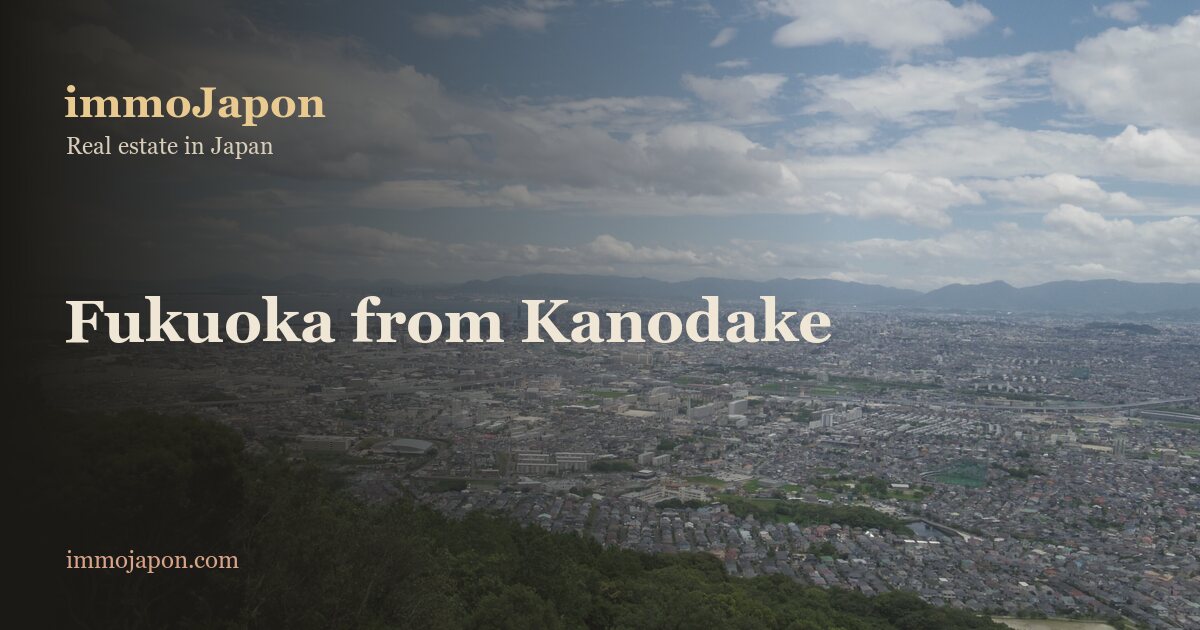 View over Fukuoka city from Mount Kanodake in Nishi ward: wooded hills in the foreground, then an unbroken fabric of low detached houses and small apartment blocks stretching to Hakata Bay under a summer sky