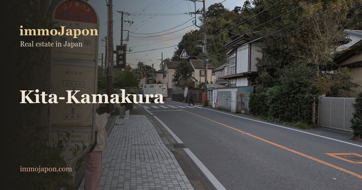 A residential street in Yamanouchi, Kita-Kamakura: a yellow bus stop sign for Kamakura station, a paved footway along a narrow road, low Japanese detached houses behind clipped hedges and, in the background, the wooded hills that close in the valley