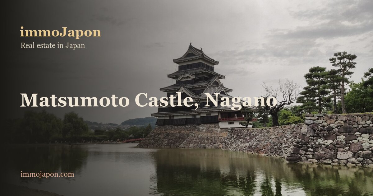The black five-storey main keep of Matsumoto Castle in Nagano Prefecture, one of Japan's twelve original castle keeps, rising above its stone base and the calm water of the moat under a grey sky, with trees along the bank.