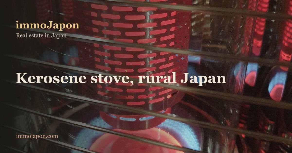 Close-up of a Japanese kerosene stove burning in a rural house: a chrome guard of horizontal bars in front of a perforated cylinder glowing bright red, a ring of blue flame around the circular burner plate at its base, flame reflections on the metal walls, and the maker's plate along the bottom rail