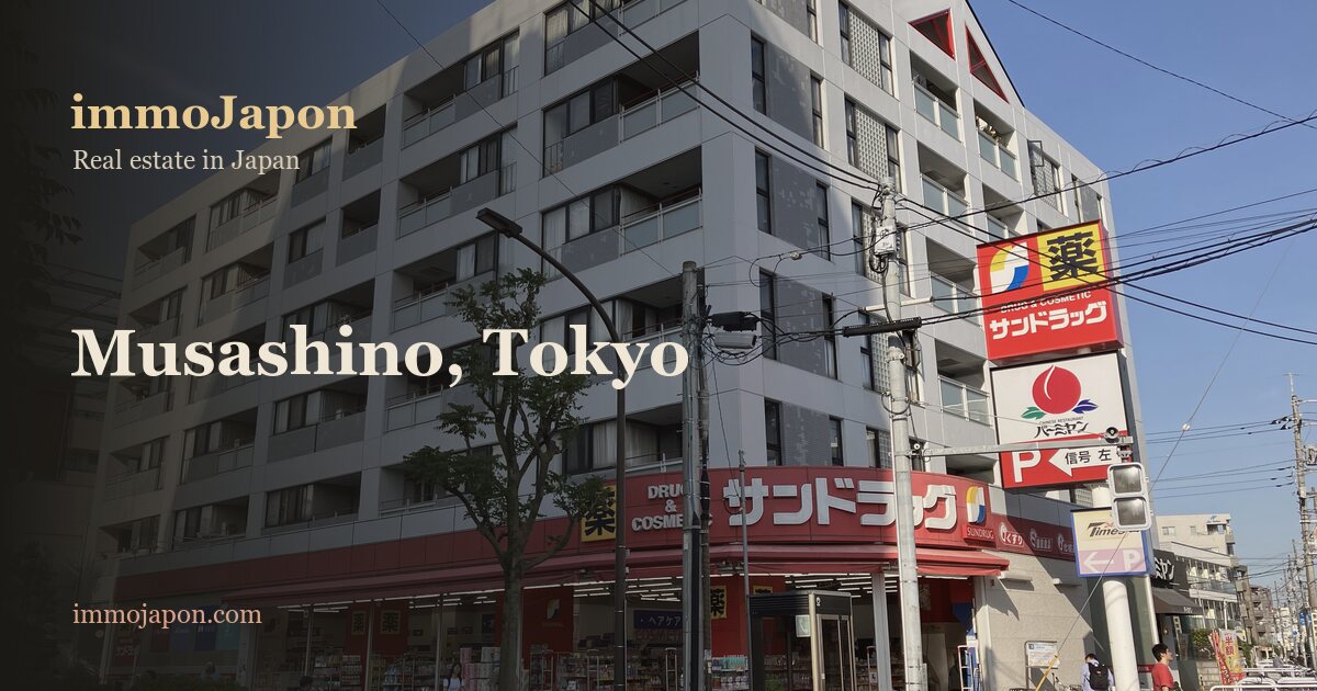 An ordinary street in Midorichō, Musashino, in Tokyo's western suburbs: a white six storey apartment block sitting above a neighbourhood pharmacy, balconies and air conditioning units on the facade, overhead power lines, traffic lights and a pedestrian crossing, the kind of rental housing a newcomer to Japan moves into