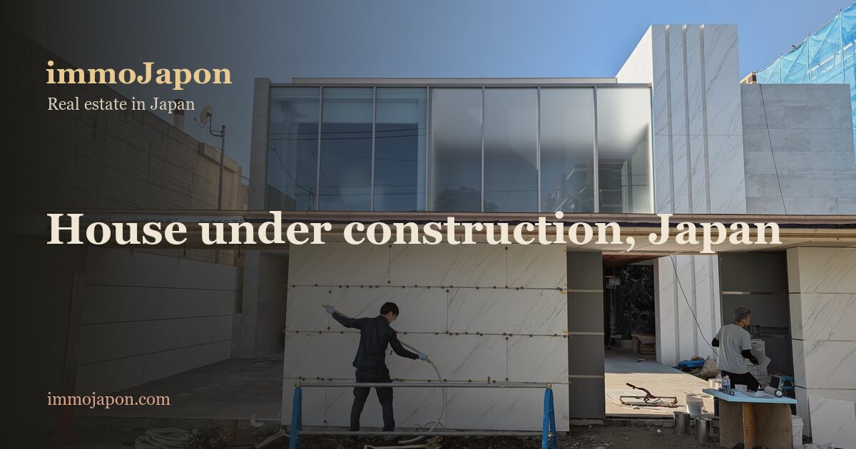 Japanese house under construction on a clear blue day: two workers fit large pale stone facade panels onto the ground-floor frame, a full-width glazed bay runs across the upper storey, and blue trestles, an orange cone, hoses and blue site netting fill the foreground