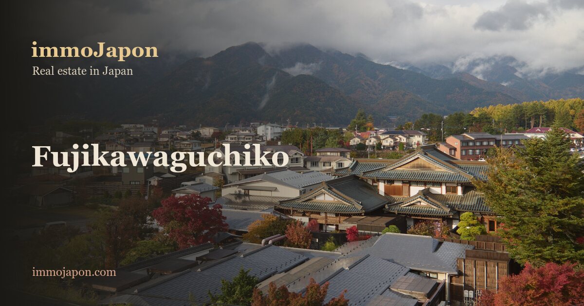 A raised view over the town of Fujikawaguchiko at the foot of Mount Fuji: Japanese detached houses with dark tiled roofs, air-conditioning units on the facades, cars parked along a residential street, red autumn maples and cloud-covered mountains behind, the ordinary fabric where a homeowner's budget is actually spent