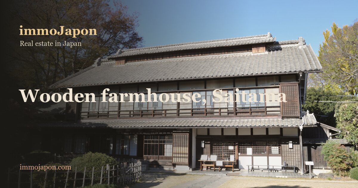 A large two-storey Japanese wooden farmhouse with layered grey tiled roofs, a band of glazed sliding windows along the upper floor under a deep eave, dark timber posts and white plaster panels on the ground floor, wooden shutters at each end, a low bamboo fence and clipped hedges on the left, a gravel forecourt in the foreground, russet maples and a clear blue autumn sky