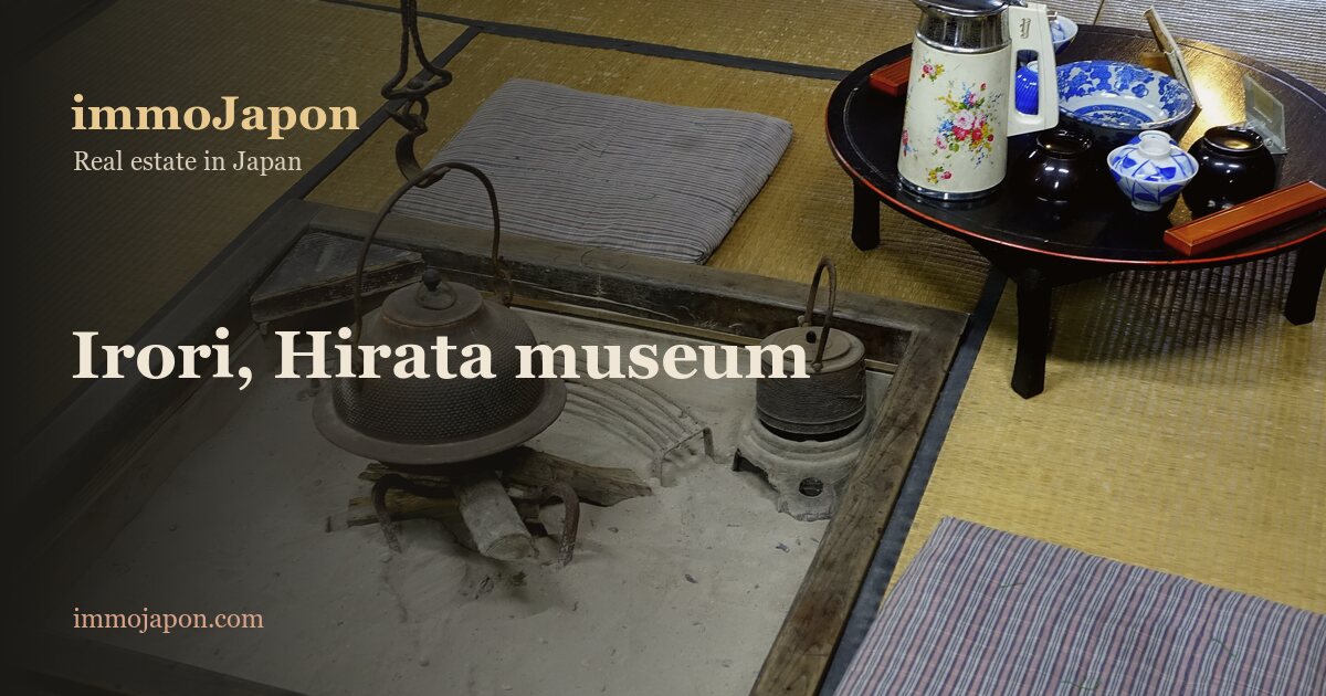 An irori hearth set into the floor of a merchant house in Takayama: a square of grey ash edged with a dark timber frame, a few logs at its centre, a lidded cast-iron pot hanging from a hook above the fire, a small kettle on a stand to the right, and a grey cushion and a lacquered low table holding a teapot and blue and white bowls on the tatami around it