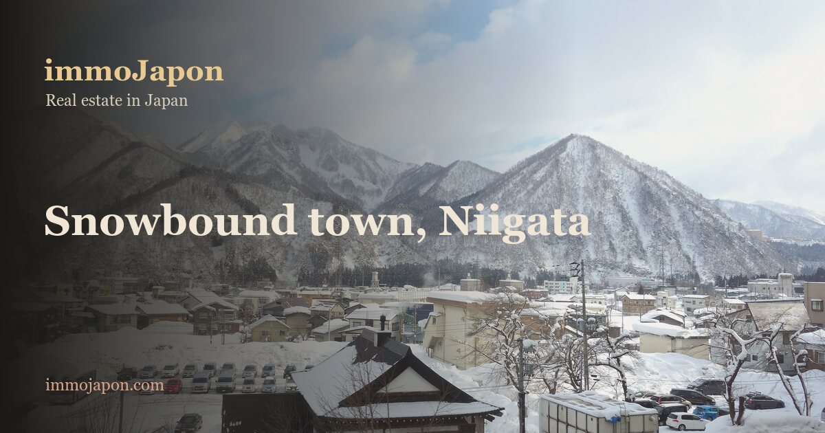 Snow covered town of Yuzawa in Niigata prefecture seen from above: two and three storey houses whose roofs and streets have disappeared under deep snow, a car park cleared between high snow banks, a traditional dark timber house in the foreground, and a ridge of snow covered mountains closing the valley under a pale winter sky.
