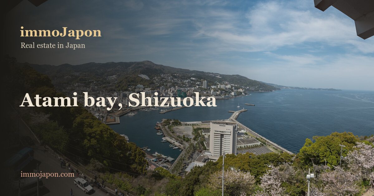 A high view over Atami bay from the castle: the hot spring resort terraced along the hillside, its white buildings and tight roofs running down to the marina and breakwater, blue sea filling the right half of the frame, cherry blossom in the foreground and the tiled eaves of the keep across the top of the picture