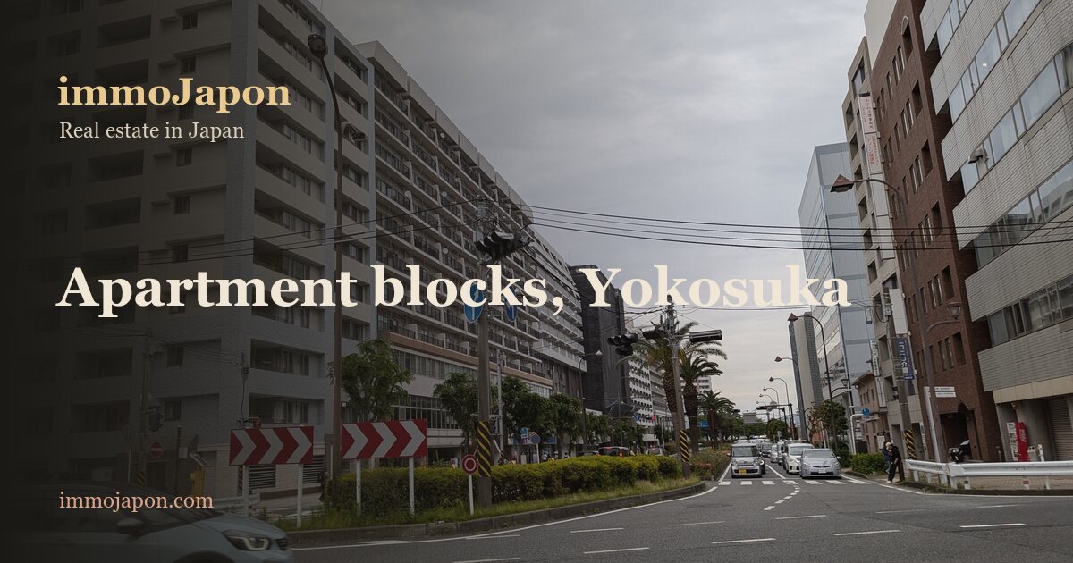 A wide avenue in central Yokosuka lined with a long ten-storey apartment block whose balconies run the length of the facade, a crossroads with painted lane markings in the foreground and a pale blue car, palm trees and a clipped hedge on the median, traffic lights and red and white chevron signs, under an overcast sky