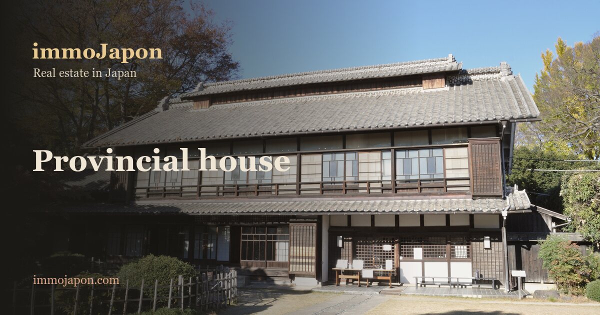 Large two-storey Japanese country house in Fukaya, Saitama prefecture: dark timber and white plaster facade, grey kawara tiled roof, clipped hedge and gravel forecourt under a blue sky, an example of the rural houses foreign buyers acquire