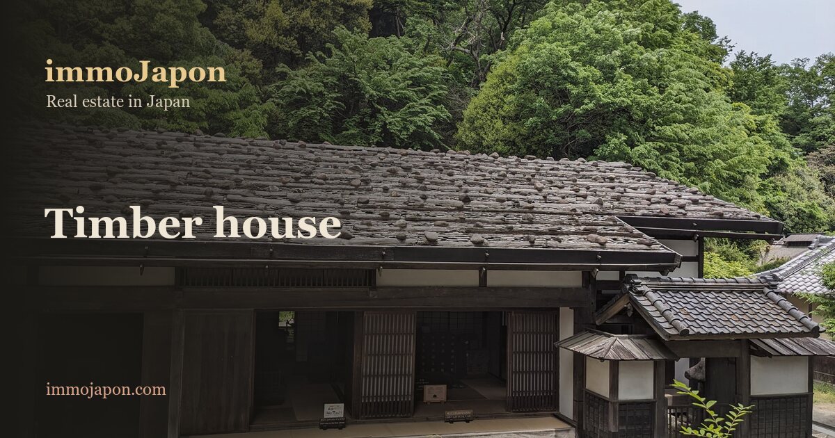 An old Japanese timber house: a long shingled roof weighted down with stones, deep overhanging eaves, dark boarded walls with wooden sliding doors, a tiled porch roof to one side and a wooded hillside behind, the kind of traditional building a buyer arriving from Europe discovers