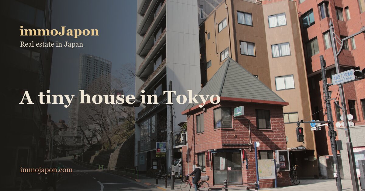 A small two-storey red-brick house with a steep dark gable roof standing on a street corner near Roppongi in Tokyo, squeezed between much taller apartment and office buildings, with a pedestrian crossing and traffic lights in the foreground.