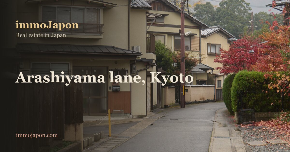 A residential lane in Arashiyama, Kyoto, in autumn: two-storey houses with grey tiled roofs and wooden shutters, boundary walls, a red maple on the right and a utility pole under a hazy sky